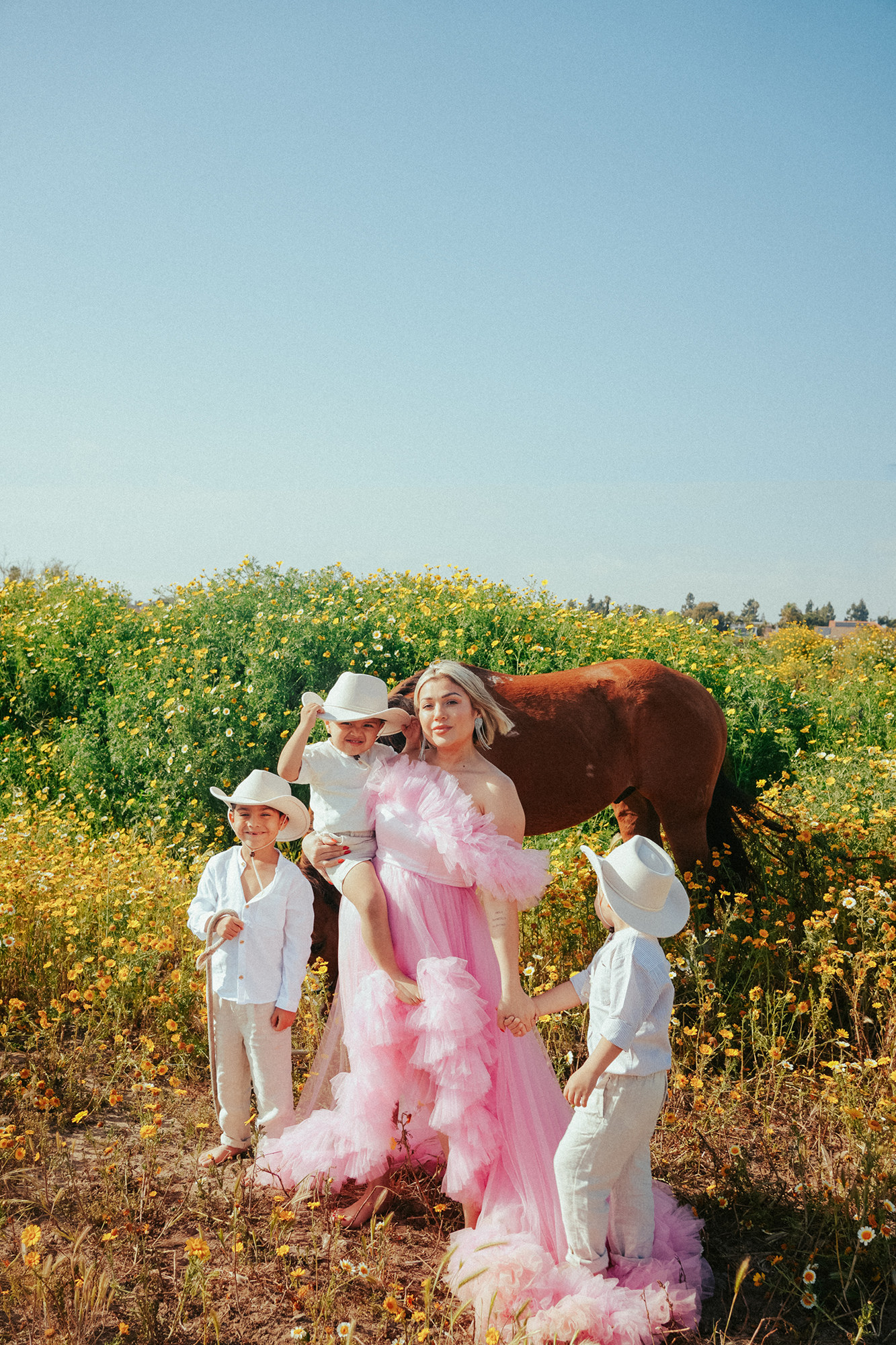 woman with short blonde bob haircut wearing a pink tulle dress standing in a field of yellow flowers with her three sons wearign coyboy hats and a horse standing behind them in San Diego